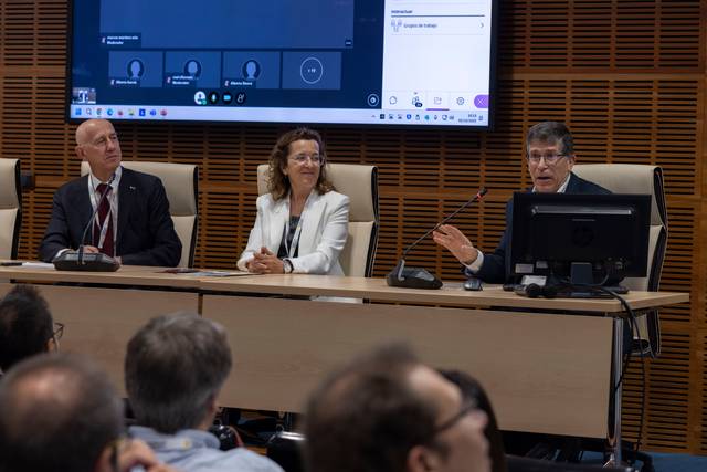 Una mesa de conferencias con tres personas sentadas frente a una pantalla que muestra una videoconferencia.