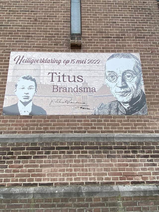 A mural featuring two images of Titus Brandsma and text, mounted on a brick wall.