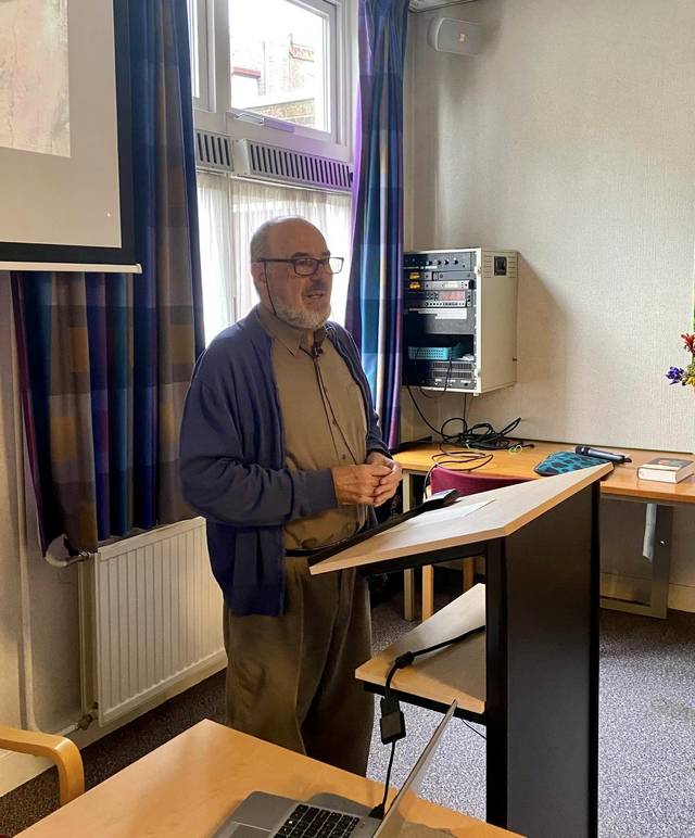 A man standing in a classroom beside a podium, with a computer and printer in the background.