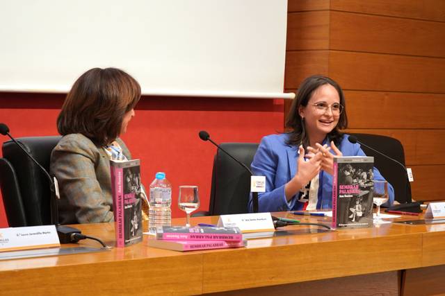 Dos mujeres participan en un evento, una de ellas hablando con gestos animados mientras la otra escucha.