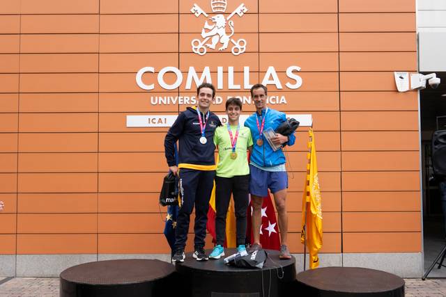 Three athletes stand on a podium outside the Comillas University building, displaying their medals.