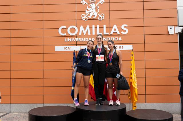 Three athletes standing on a podium in front of the Comillas Pontifical University, holding medals and flags.