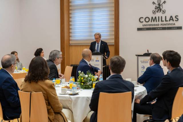 Un grupo de personas se sienta alrededor de una mesa en un evento en la Universidad Pontificia Comillas, mientras un orador se dirige a ellos.