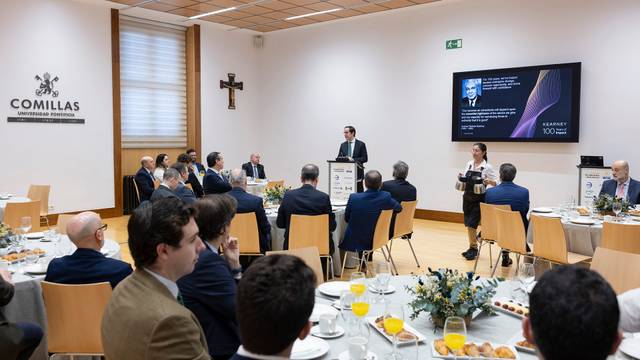 Una reunión formal en un comedor con varias personas escuchando a un conferencista frente a una presentación en pantalla.