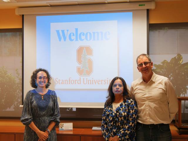 Tres personas posan frente a una presentación que da la bienvenida a la Universidad de Stanford.