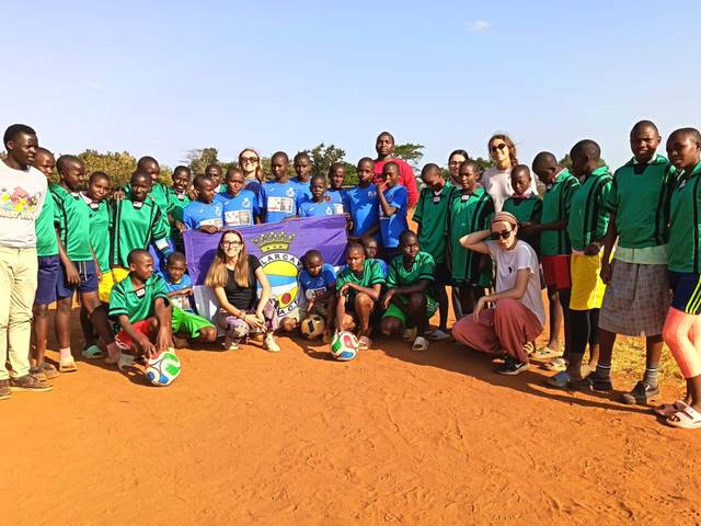 Un grupo de niños y adultos posa en una cancha de tierra con balones de fútbol y una bandera.