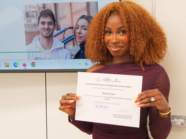 Una mujer sonriente sostiene un certificado frente a una pantalla que muestra a dos personas.