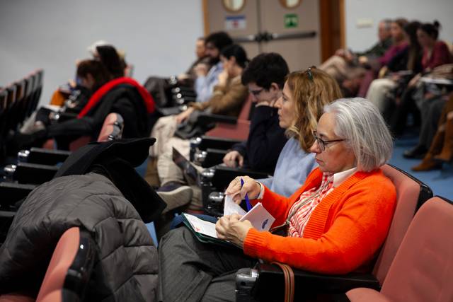 Una audiencia atenta en una sala de conferencias, algunos asistentes usan dispositivos electrónicos.