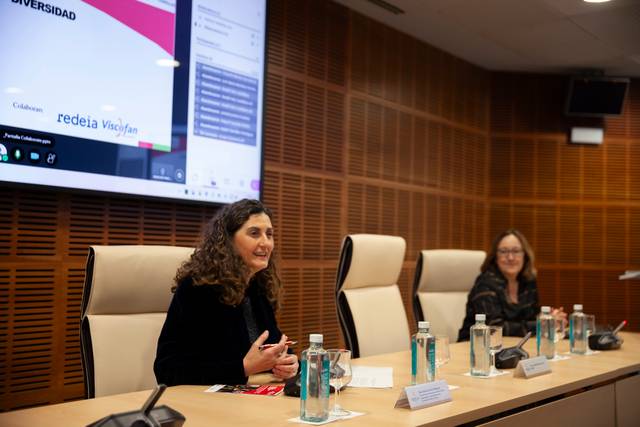 Una mujer habla en una mesa durante un evento, mientras otra mujer la escucha en un entorno académico.