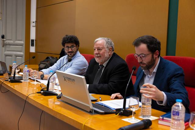 Una mesa de debate con tres hombres sentados frente a una audiencia, hablando sobre un tema de interés.