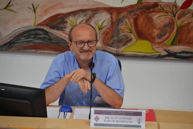 A man with glasses sitting at a desk with a microphone, behind a nameplate in a room with a colorful mural.