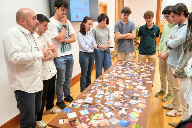 Un grupo de jóvenes observa atentamente una mesa llena de fotos y tarjetas mientras un hablante les explica algo.