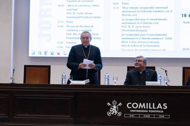 Imagen de un evento académico en el que dos sacerdotes están sentados en una mesa frente a una presentación.