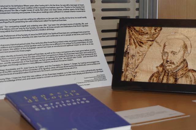 La imagen muestra un cuadro con un retrato de un hombre, un libro sobre la mesa y un documento enmarcado con texto.