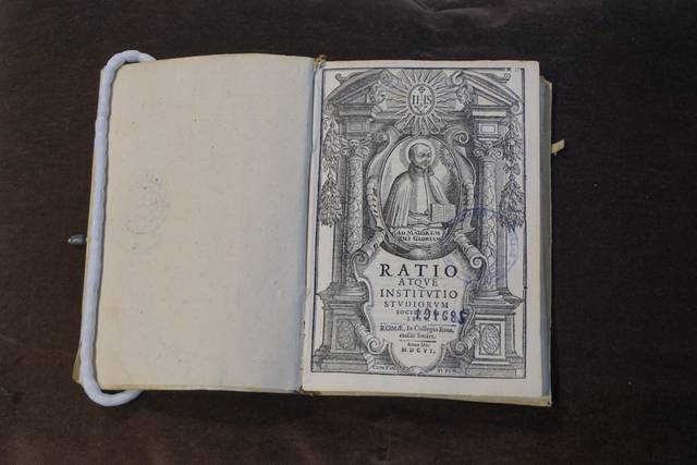 El libro abierto muestra una página con un grabado y el título 'RATIO ATQUE INSTITUTIO STUDIORUM'.