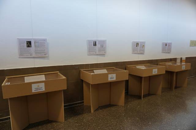 La imagen muestra una exposición con vitrinas de cartón y paneles informativos en la pared.