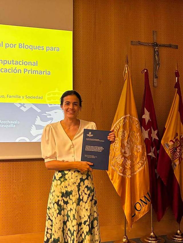 Una mujer sonriente sostiene un documento en un entorno académico con banderas y una cruz al fondo.