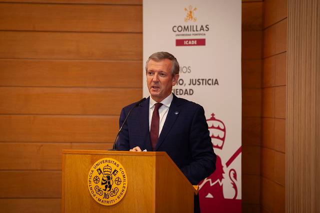 A man in a suit is giving a speech at a podium with a university emblem and a backdrop with text.