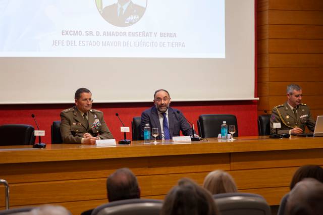 Un grupo de personas en una mesa durante un evento oficial, con un fondo que muestra un retrato y una leyenda.