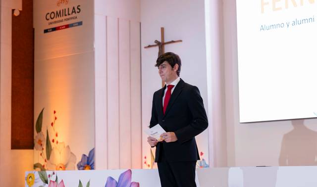 Un estudiante de pie frente a un auditorio, sosteniendo un papel y vestido formalmente con traje y corbata.