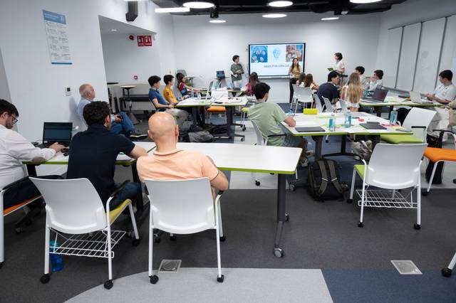 Una sala de clases moderna con estudiantes sentados en escritorios mientras un grupo presenta en una pantalla.