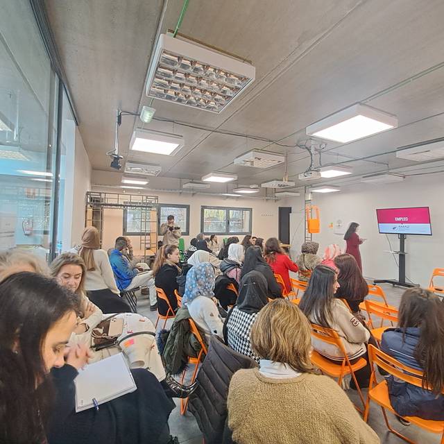 Una sala llena de mujeres sentadas en sillas naranjas, prestando atención a una presentación en una pantalla.