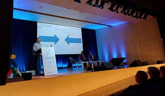 Un presentador habla en un evento mientras otros participantes lo escuchan en un escenario con iluminación azul.