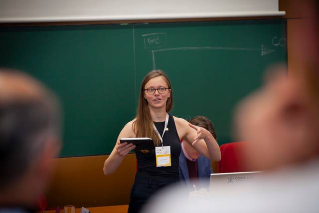 A woman stands at the front of a classroom, speaking to an audience while holding a tablet.