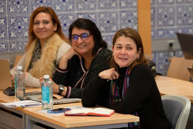 Tres mujeres sonrientes sentadas en un salón de clases con libros y botellas de agua en la mesa.