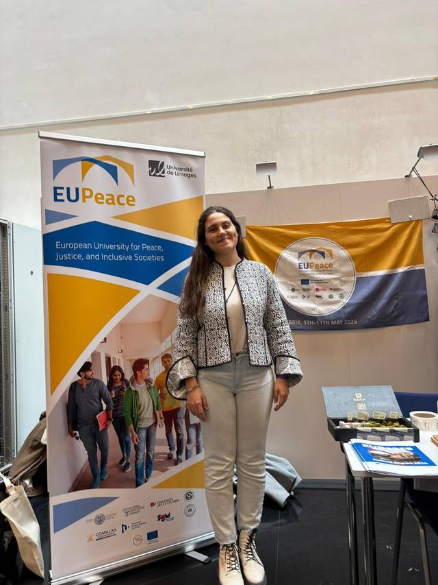 Una mujer sonriente se presenta en un evento junto a un banner de la Universidad Europea para la Paz, la Justicia y las Sociedades Inclusivas.