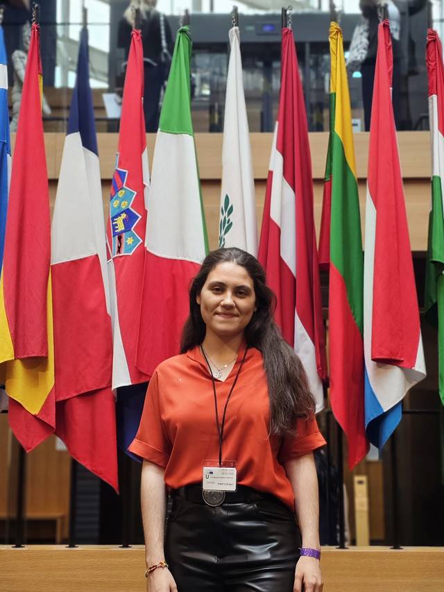 Una mujer sonriente se encuentra frente a una serie de banderas de diferentes países en un edificio institucional.