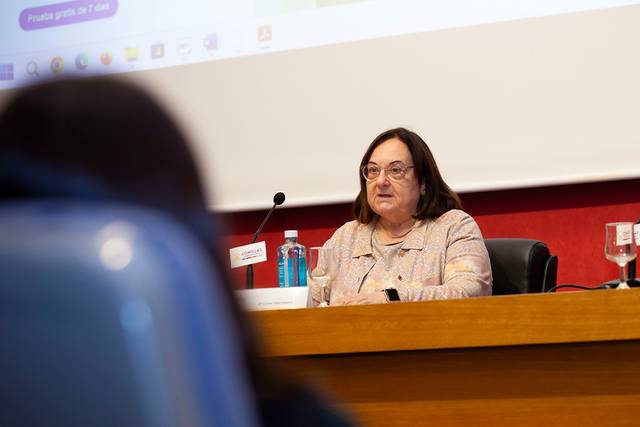 Una mujer está sentada detrás de una mesa hablando en un micrófono durante un evento en una sala con audiencia.