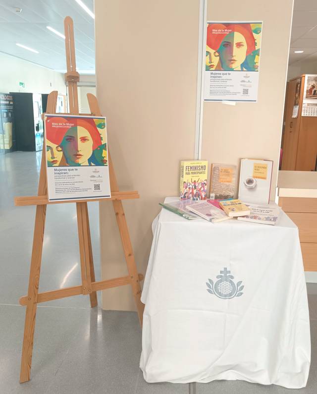 La imagen muestra un caballete con carteles y una mesa cubierta con una mantelería blanca que exhibe libros sobre feminismo.