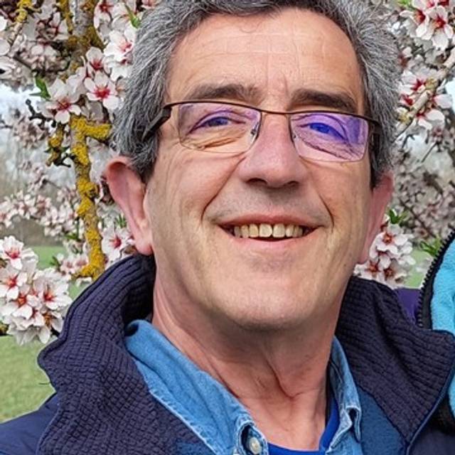 An older man with glasses smiling in front of blooming cherry blossoms.