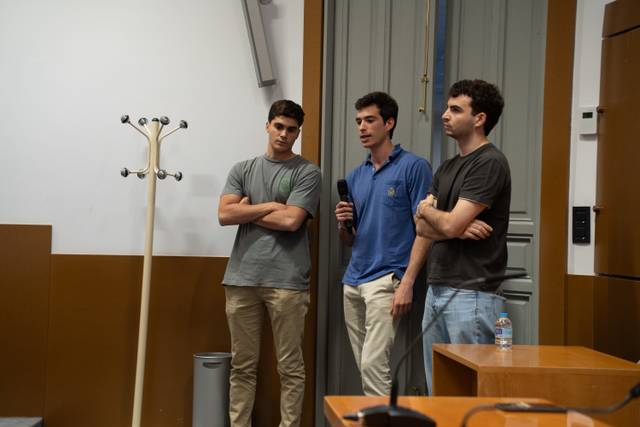 Tres jóvenes están de pie frente a una audiencia mientras uno de ellos habla.
