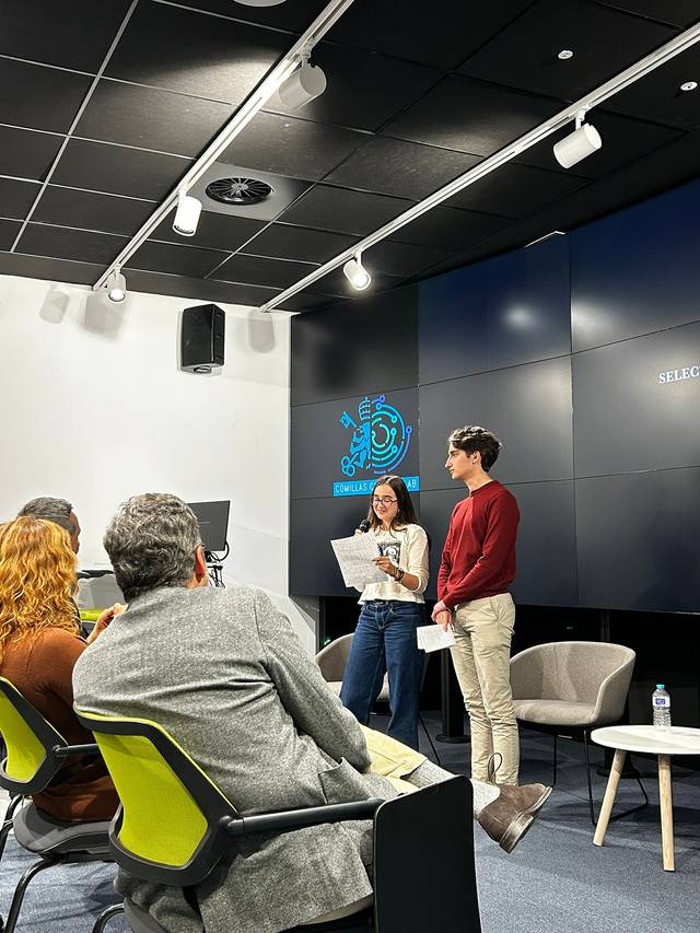 Dos presentadores están hablando ante un público en un entorno de oficina moderno.