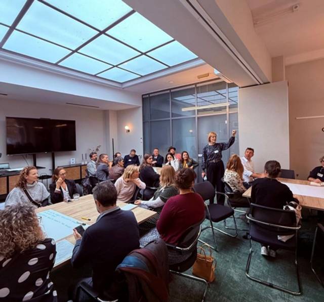 Una reunión en un espacio moderno con varios participantes atentos y un presentador en primer plano.