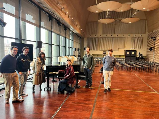 Un grupo de seis personas posando en un amplio salón con un piano y muchas sillas vacías.