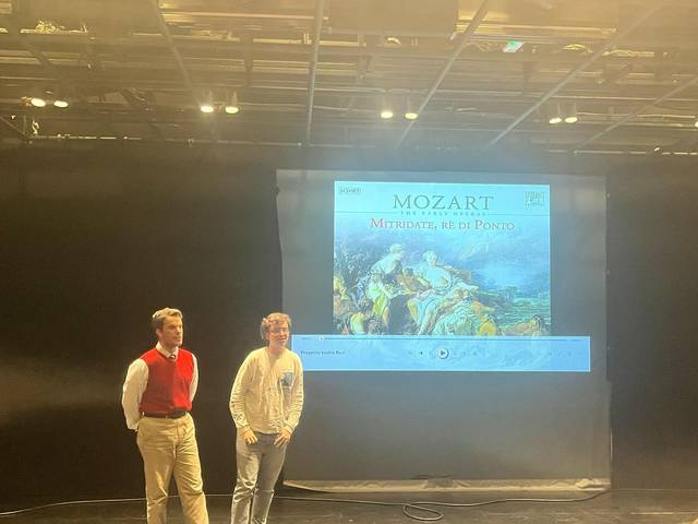 Dos personas de pie frente a una proyección que muestra un cartel de una obra de Mozart.
