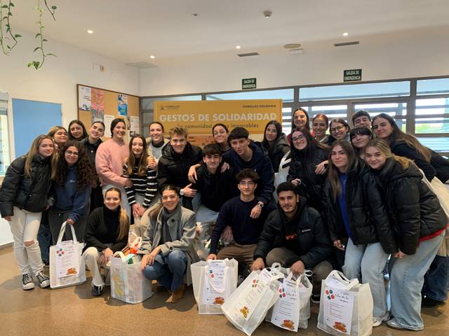 Un grupo de jóvenes sonríen para la foto en un ambiente interior, posiblemente en una actividad de solidaridad, rodeados de bolsas de comida.