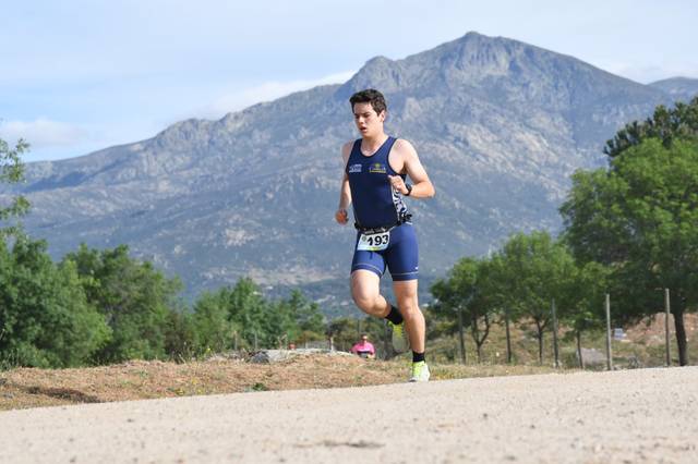 A young male athlete in a blue triathlon suit is running on a dirt path with a mountainous background under a clear sky.