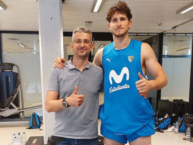 Two men, one taller and in a blue sports uniform, standing in an indoor setting giving thumbs up.