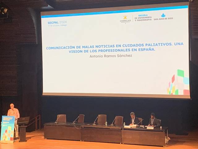 A person speaking at a conference podium with a presentation slide about palliative care communication in Spain, while several attendees sit onstage.