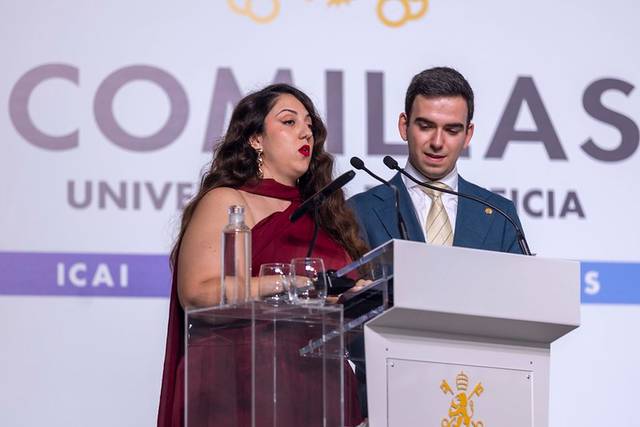 Dos personas en un evento formal están hablando desde un podio frente a una pantalla que muestra el logotipo de la universidad.