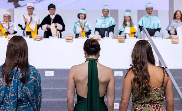 Tres mujeres de espaldas observan un evento académico con graduados y autoridades en el escenario.