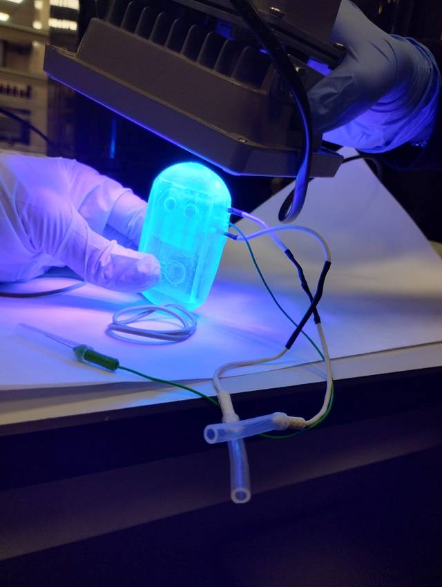 Un dispositivo iluminado en azul está siendo sostenido por una mano con guantes, conectado a unos cables.