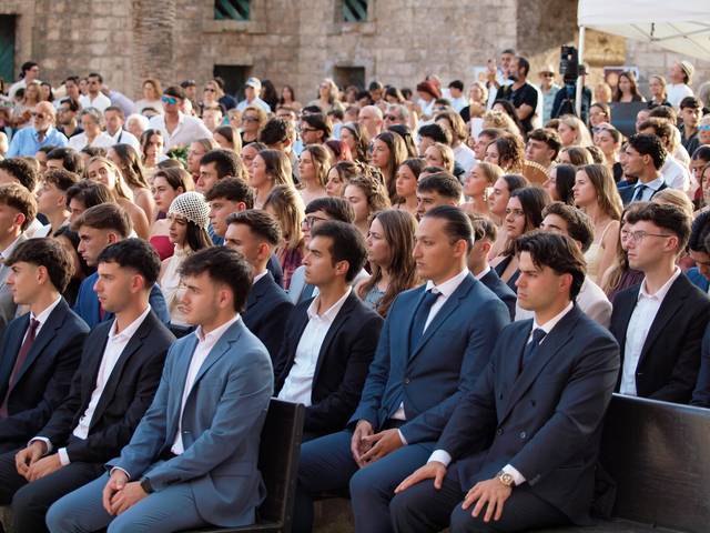 Una multitud de jóvenes sentados en un evento al aire libre, algunos vestidos formalmente.