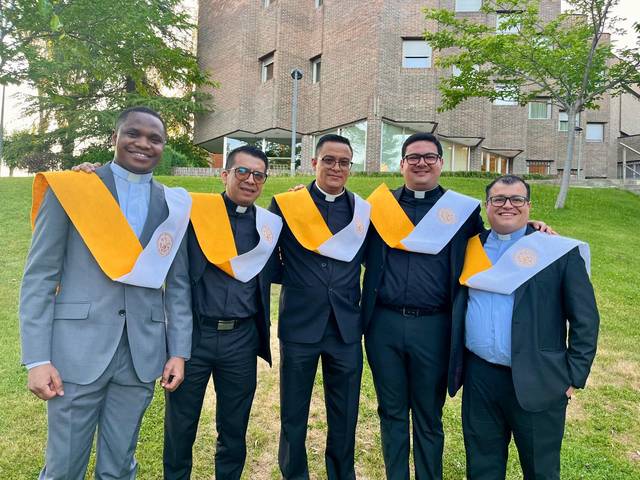 Un grupo de cinco hombres vestidos con trajes formales y togas de graduación posan juntos al aire libre.