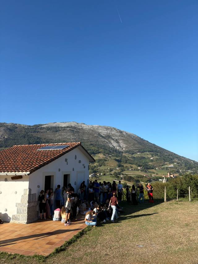 Un grupo de personas se congrega frente a una casa blanca en un paisaje montañoso bajo un cielo despejado.