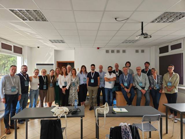 A group of people, mostly standing, posing for a photo in a classroom with desks and a whiteboard.